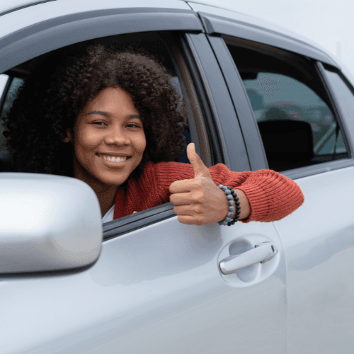 Smiling person sitting in a car, giving a thumbs up through the open window.